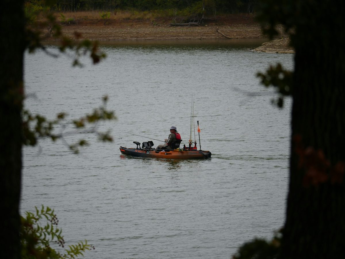 Fully rigged fishing kayak with a safety flag on a lake, framed through trees